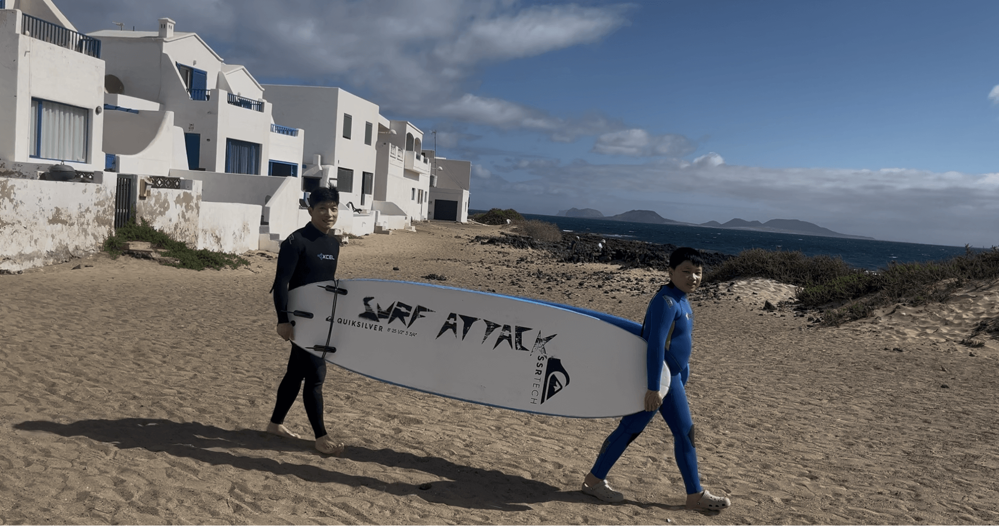Two surfers in wetsuits carrying a Surf Attack surfboard on Famara Beach, Lanzarote
