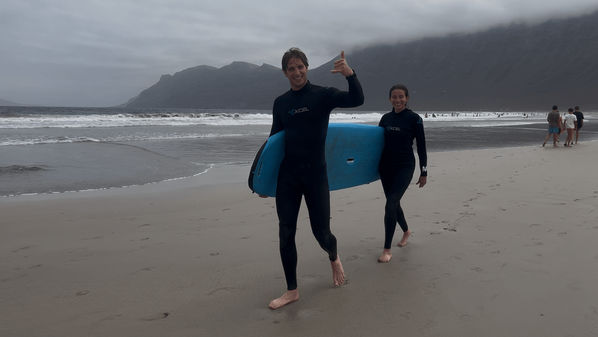 Two surfers in wetsuits carrying a blue surfboard on Famara Beach during a private surf lesson in Lanzarote