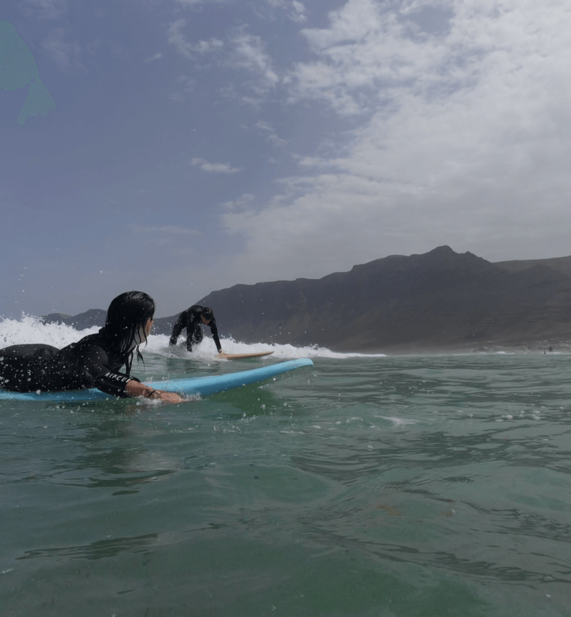 Two surfers taking a private surf lesson on Famara Beach, Lanzarote with mountainous background and cloudy sky